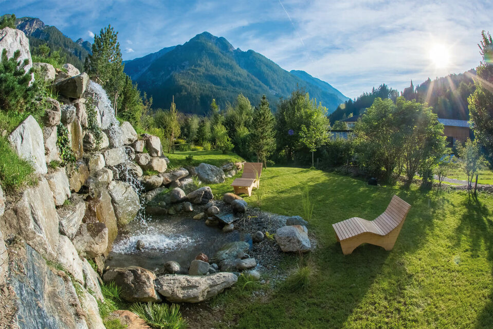 Zwei Zirbenliegen laden im Schaugarten ‚Vitalpinum‘ in Thal zum Entspannen ein, während ein kleiner Wasserfall davor beruhigend über die Steine plätschert.
