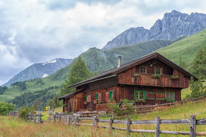 Das Bild zeigt die Gottschaunalm, eine Almhütte aus Holz auf einer Bergwiese. Ein Holzzaun verläuft entlang des Hanges vor dem Gebäude, während im Hintergrund grüne Berghänge und markante Felsgipfel zu sehen sind. Die Hütte liegt in ruhiger Lage inmitten der alpinen Berglandschaft.