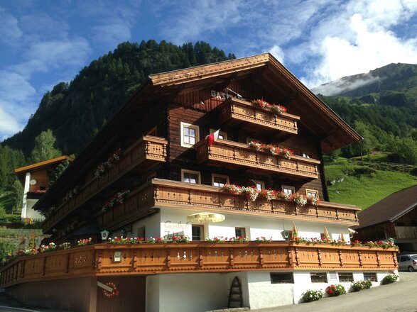 Das Bild zeigt den Gasthof Islitzer, ein mehrstöckiges Gebäude in traditioneller Holzbauweise mit zahlreichen Balkonen. Die Balkone sind mit Blumenkästen geschmückt, und im Erdgeschoss befindet sich eine große Sonnenterrasse mit Holzgeländer. Im Hintergrund sind grüne Wiesen und bewaldete Berghänge zu sehen, die die alpine Lage des Gasthofs unterstreichen.