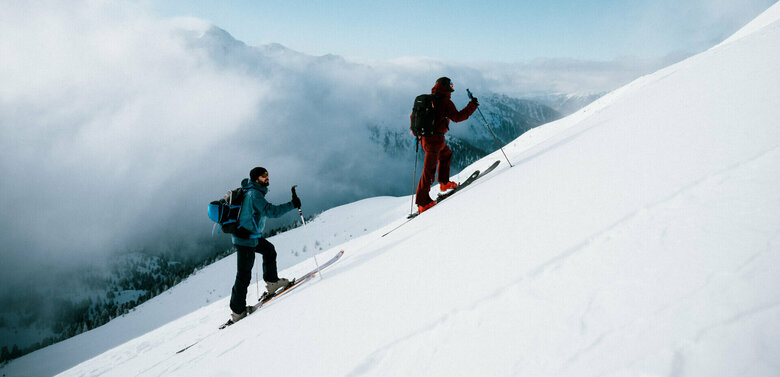 Zwei Skitourensportler erklimmen im Gleichschritt auf ihren Tourenskiern einen mittelsteilen Berghang an einem Schönwettermorgen. Dahinter verdeckt zäher Hochnebel einen Teil der Berge.
