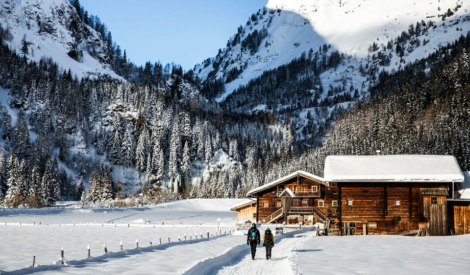 Zwei Personen spazieren auf einem Winterwanderweg an einer kleinen Ansammlung von kleinen Hütten vorbei. Dahinter der bewaldete Talschluss in einer winterlichen Umgebung.