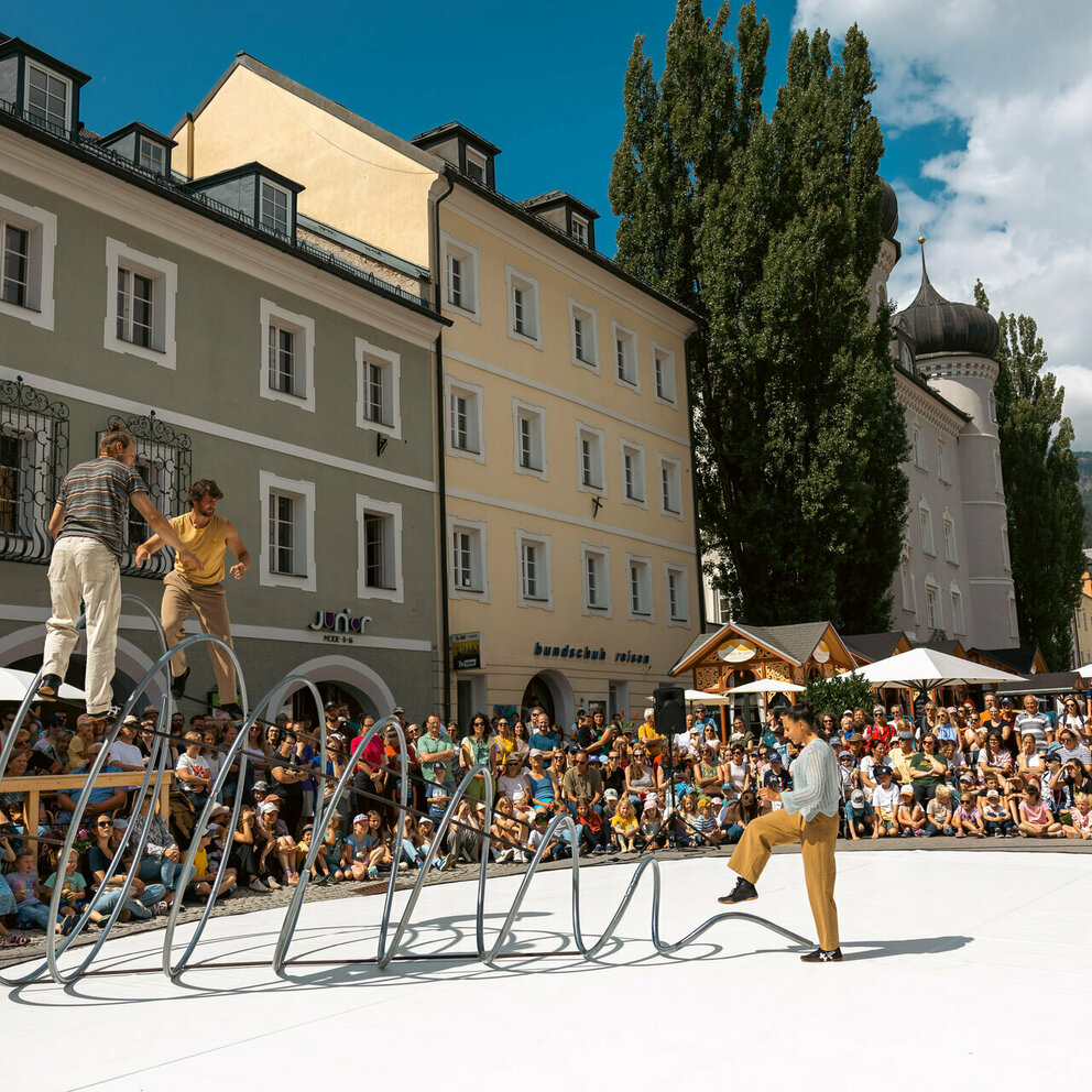 Drei Artisten turnen auf, in bzw. vor einer großen Spirale aus Eisen am Lienzer Hauptplatz im Rahmen von Olala 2025. Sie werden vom begeisterten Publikum rundherum bestaunt.