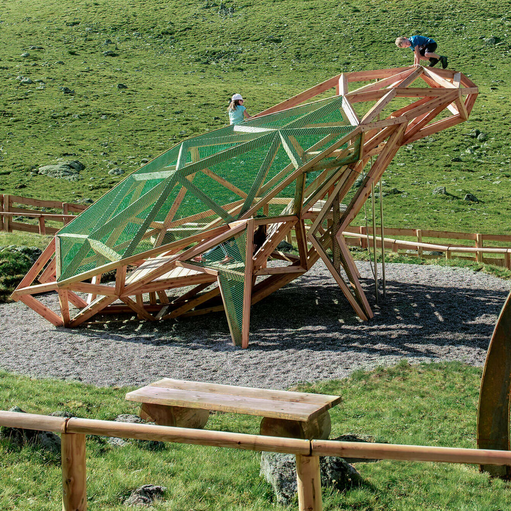 Kinder spielen auf einem überdimensional-großen Frosch, einem der vielen Spielgeräte des Wassermythos Ochsenlacke im Skizentrum St. Jakob i. D.. Die Umgebung außerhalb des eingezäunten Spielgerätes ist eine saftig grüne Wiese.
