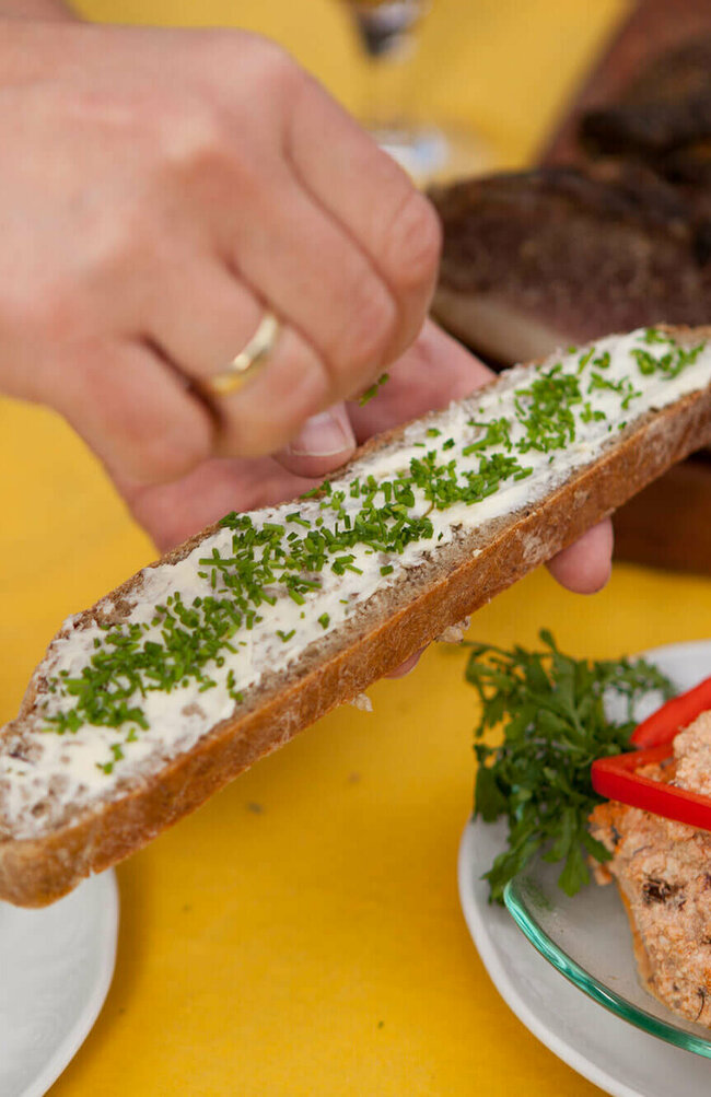 Eine Scheibe Vinschgauer Brot mit Butter und Schnittlauch. Daneben ein weißer und ein roter Aufstrich und im Hintergrund Speck.