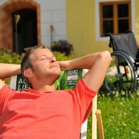 Ein Mann liegt in einem Liegestuhl und genießt die Sonne. Im Hintergrund steht ein Rollstuhl.