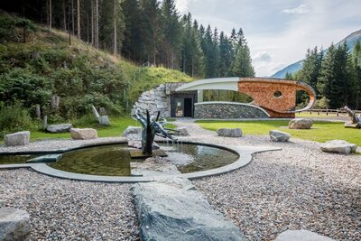 Der Außenbereich der Deferegger Heilwasserwelt in St. Jakob in Defereggen. Der Brunnen gefüllt mit Heilwasser ist der Mittelpunkt des Bildes, dieser ist umgeben von Kieselsteinen. Im Hintergrund ist die sogenannte "Serona-Höhle" zu sehen, welche von grünem Gras und einem dichten Wald umgeben ist.