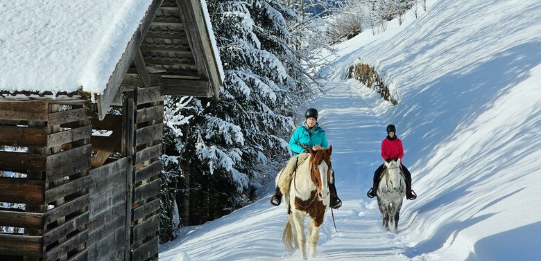 Zwei Frauen reiten mit einem braun-weißen und einem grauen Pferd auf einem schneebedeckten Weg in Prägraten. Links steht eine schneebedeckte, hölzerne Hütte.