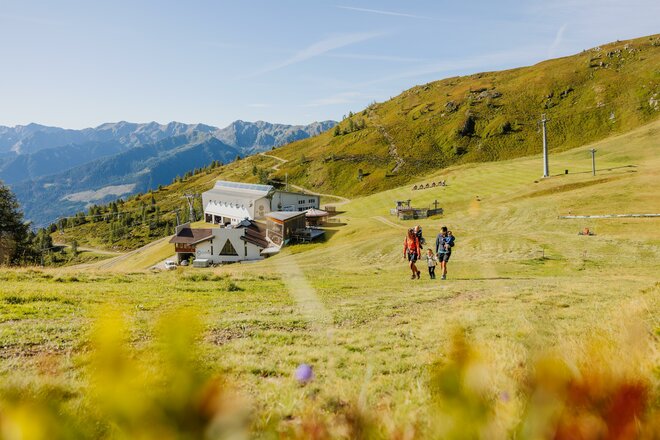 Drei Menschen spazieren durch ein Feld mit Rucksäcken. Im Hintergrund sieht man die Gondelstation und ein Gebäude.