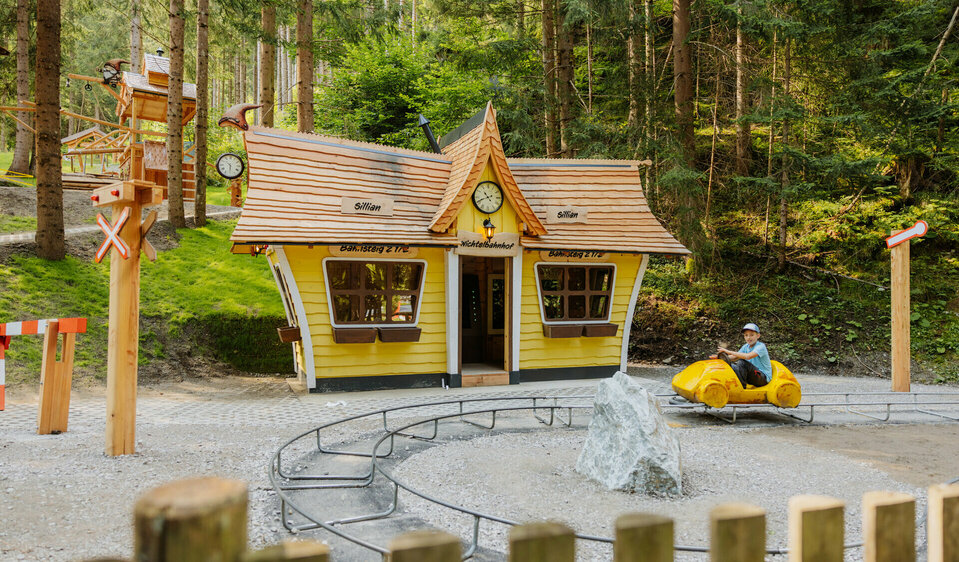 Auf dem Bild ist der neue Bahnhof des Wichteldorfes zu sehen, vor dem ein kleiner Junge mit einem  Miniatur Tretauto vor dem Bahnhof seine Runden dreht.