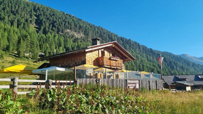 Hinterpasslerhütte aus Holz mit Sonnenschirmen vor bewaldetem Berghang unter blauem Himmel.