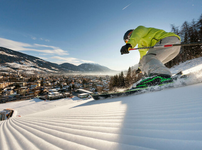 Ein Skifahrer mit gelber Jacke und heller Hose carved am Hochstein mit den Ski. Aufgenommen von unten. Blick auf das Dorf.