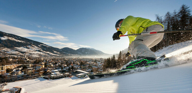 Ein Skifahrer mit gelber Jacke und heller Hose carved am Hochstein mit den Ski. Aufgenommen von unten. Blick auf das Dorf.