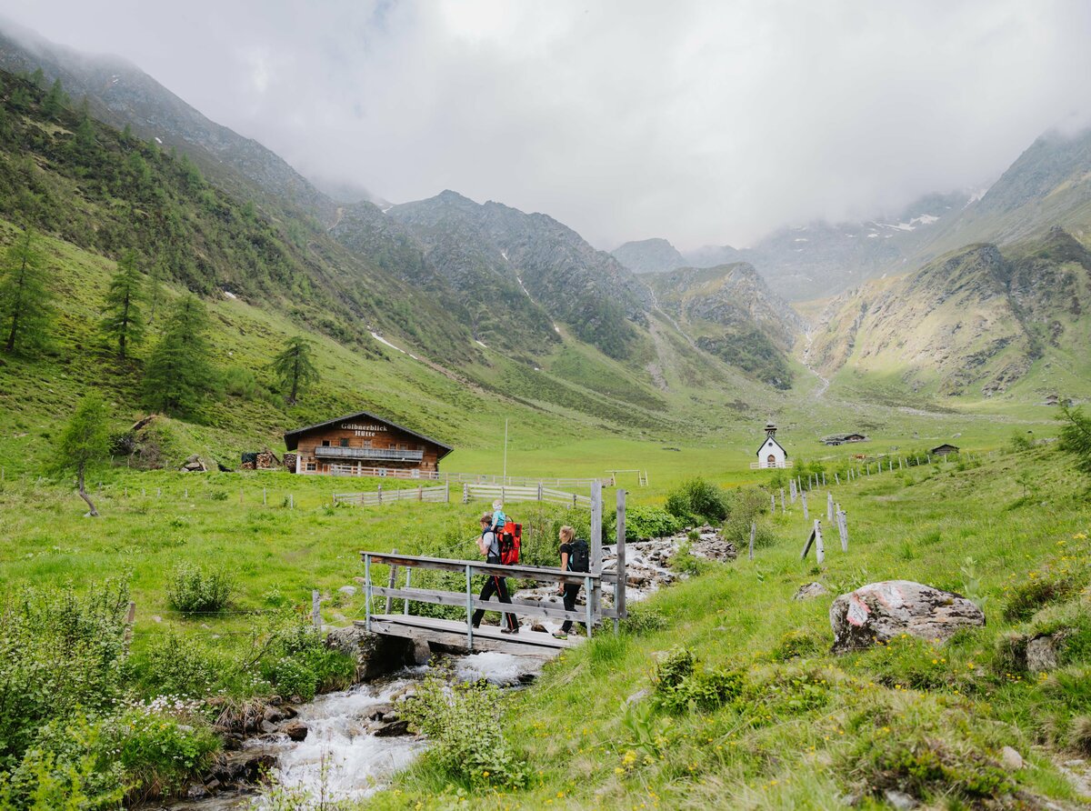 Eine junge Familie wandert über eine kleine hölzerne Brücke, über einen Bach im Kristeinertal. Sie befinden sich kurz vor der Gölbnerblickhütte. Die Berge im Hintergrund sind wolkenverhangen.