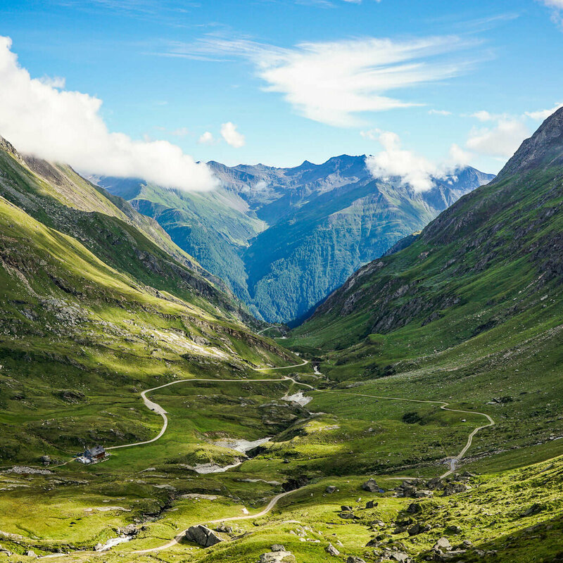 Blick auf den Adlerweg in Osttirol, Etappe 1. In einer Senke zwischen zwei Bergketten liegt eine Hütte, es fließt ein Bach und zwei Wege verlaufen entlang des Bachs. 
