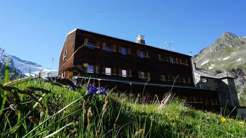 Das Bild zeigt die Essener-Rostocker Hütte mit einem mehrstöckigen Holzgebäude und einem angebauten Steingebäude. Im Vordergrund sind Gräser, einzelne Blumen und eine Bergwiese zu sehen, die bis an die Gebäude heranreicht. Im Hintergrund erheben sich bewachsene Berghänge und schneebedeckte Berggipfel unter einem klaren blauen Himmel.
