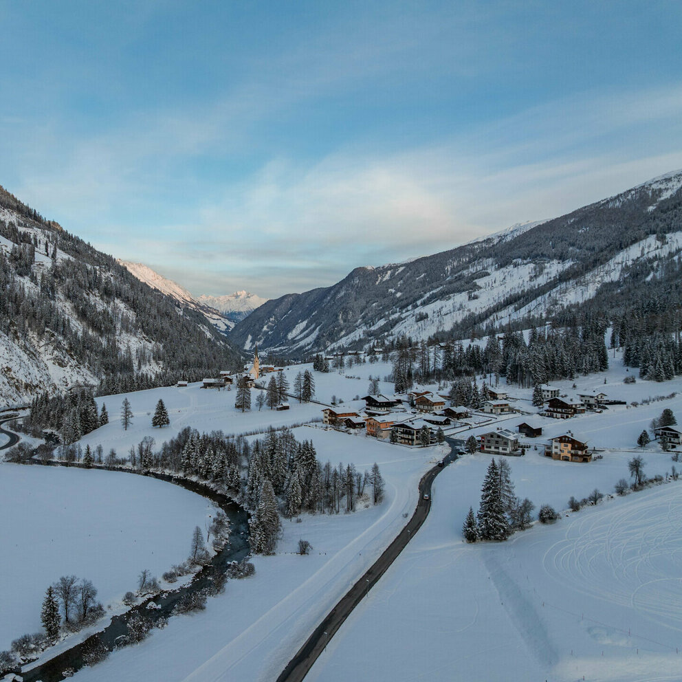 Luftaufnahme eines verschneiten Alpen­tals mit Fluss, Straßen, einem kleinen Dorf und bewaldeten Berghängen im Winterlicht.