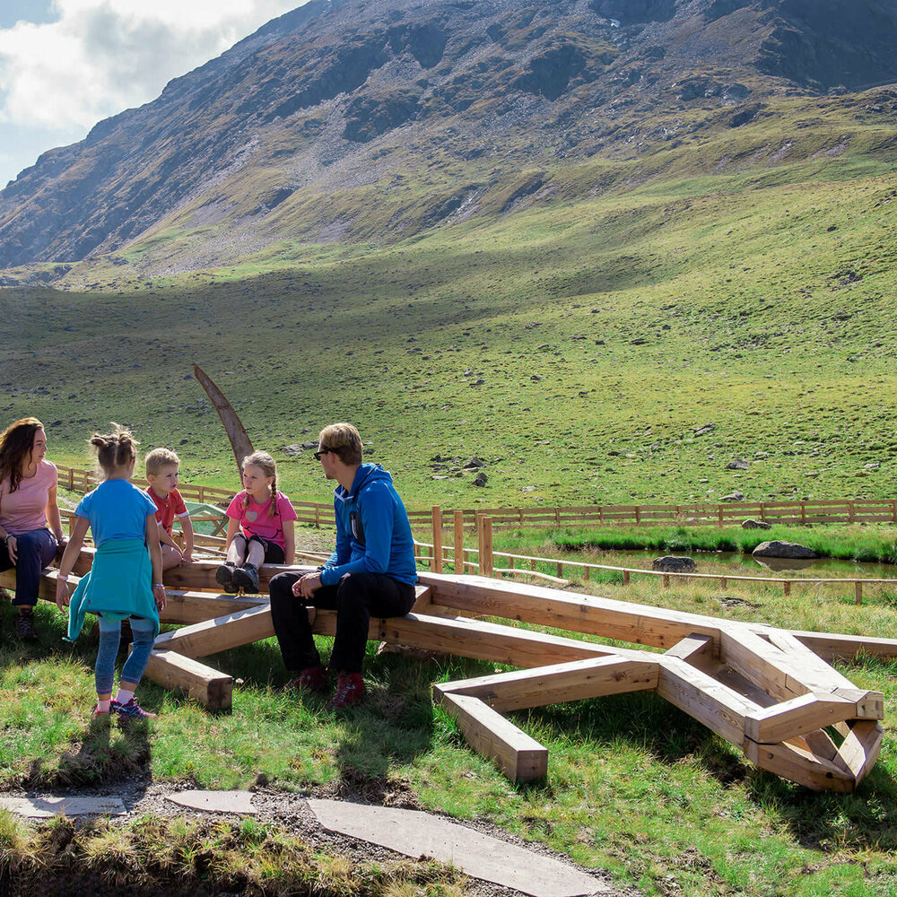 Eine Familie sitzt bei der Spielarena des Wassermythos Ochsenlacke im Skizentrum St. Jakob i. D.. Die Sonne lässt die Umgebung und die Berge im Hintergrund in einem besonderes warmen Licht erstrahlen.