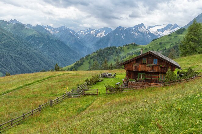 Das Bild zeigt die Gottschaunalm, eine Almhütte aus Holz auf einer weitläufigen Bergwiese. Ein Holzzaun führt über die grünen Hänge zur Hütte, während im Hintergrund zahlreiche Berggipfel und bewaldete Bergrücken zu sehen sind. Die ruhige Lage inmitten der alpinen Landschaft vermittelt einen Eindruck von Natur und Abgeschiedenheit.