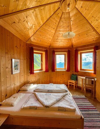 Das Bild zeigt ein Zimmer in der Neuen Sajathütte mit vollständig holzverkleideten Wänden und einer auffälligen sechseckigen Holzdecke. Im Raum stehen ein Doppelbett, ein kleiner Tisch und mehrere Stühle aus Holz. Die Fenster bieten einen weiten Blick auf die umliegende Berglandschaft und lassen viel Tageslicht in den Raum.