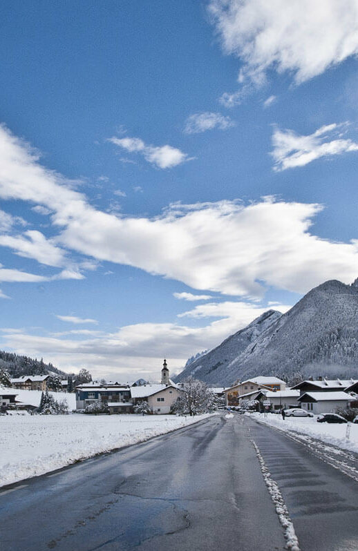 Straße, die in den Ort Abfaltersbach führt, das schon im Hintergrund zu erkennen ist. Neben der Fahrbahn liegt Schnee.