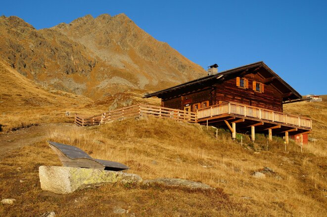 Holzhütte mit Terrasse in alpiner Graslandschaft vor einem felsigen Berggipfel unter blauem Himmel.