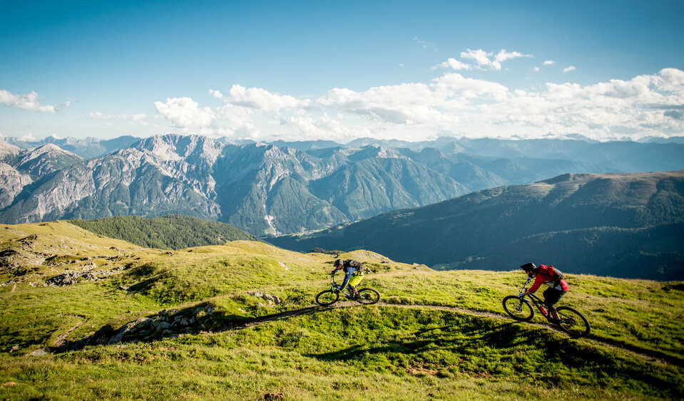 Zwei Radfahrer auf einem schmalen Trail mit einzigartigem Bergpanorama.