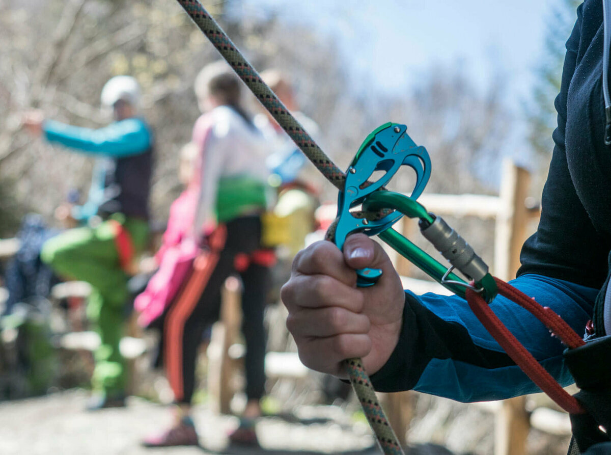 Nahaufnahme von einem Kletter-Sicherungsgerät, einem Seil und bunten Karabinern. Im Hintergrund stehen mehrere Personen.