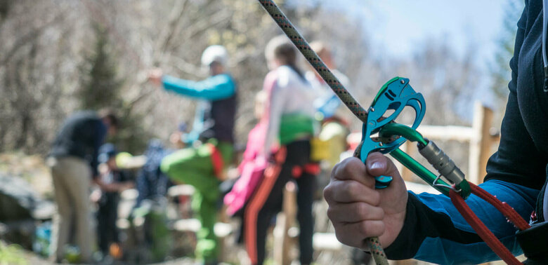 Nahaufnahme von einem Kletter-Sicherungsgerät, einem Seil und bunten Karabinern. Im Hintergrund stehen mehrere Personen.