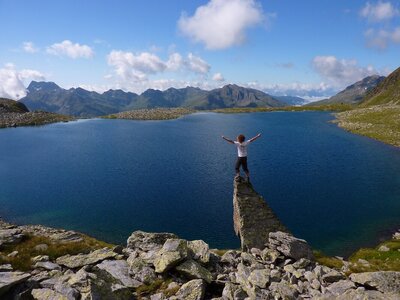 Blick auf den tiefblauen Oberseitsee unter der Seespitze in St. Jakob i. D.. Ein Mann steht auf einem Stein, der scheinbar über das Ufer hinausragt. Er streckt sich dem sommerlich-blauen Himmel entgegen.