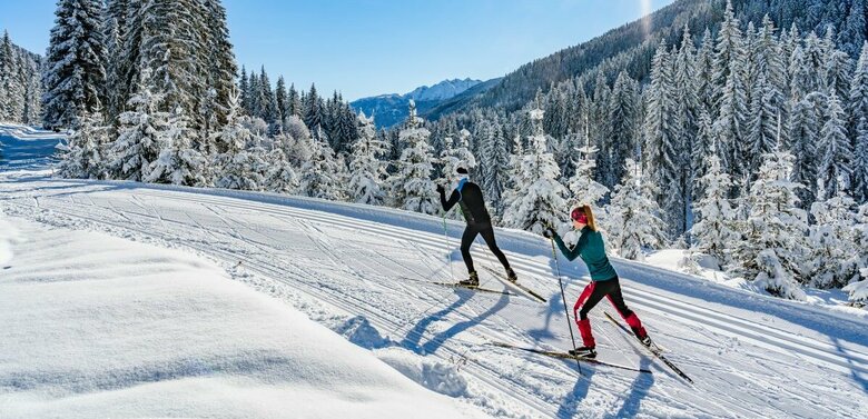 Zwei Langläufer:innen laufen bei strahlendem Sonnenschein auf der Loipe in Obertilliach mit einem frisch verschneiten Wald im Hintergrund