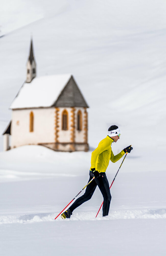Zwei Langläufer:innen laufen in den Tilliacher Feldern mit einigen kleinen Heustadeln und der Filialkirche St. Nikolaus im Hintergrund