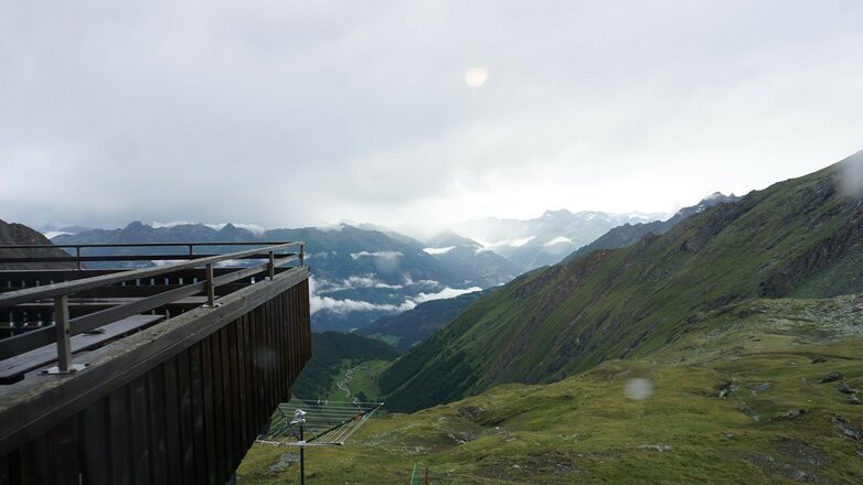 Der Blick von der Sudetendeutschen Hütte reicht über ein weites alpines Tal und die umliegenden Bergketten. Im Vordergrund sind eine Terrasse sowie grüne Berghänge zu sehen. In der Ferne liegen Wolken über den Gipfeln und Teilen des Tals.
