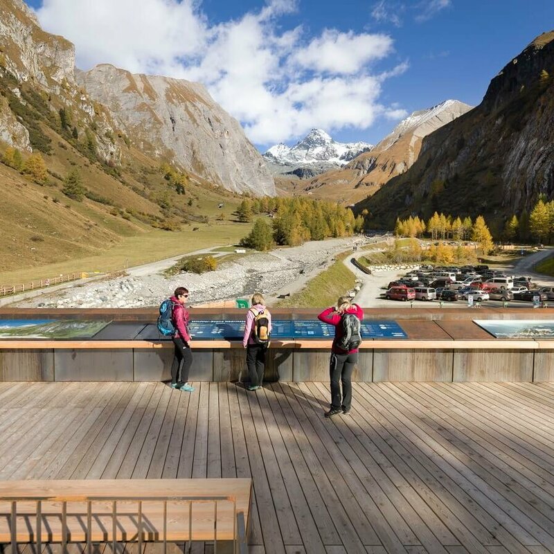 Drei Frauen stehen auf einem Parkplatz beim Glocknerwinkel in Kals und blicken auf den Großglockner. Die herbstliche Landschaft zeigt bunte Bäume und klare Sicht auf den schneebedeckten Berg.