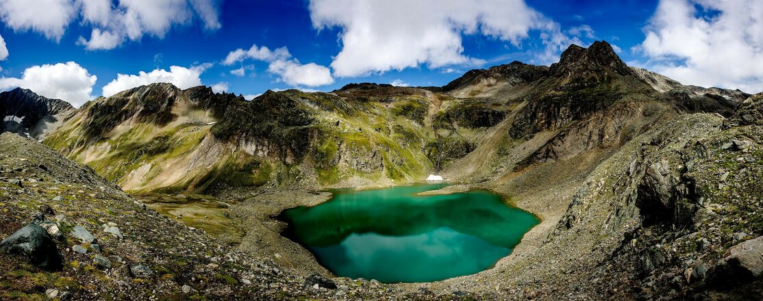 Das Bild zeigt den Eissee in einer kargen hochalpinen Berglandschaft. Der türkisfarbene Bergsee liegt in einer Mulde zwischen Felsen und Geröllfeldern und hebt sich deutlich von der umliegenden Landschaft ab. Rund um den See erheben sich felsige Berghänge und Gipfel, darüber sind weiße Wolken vor einem kräftig blauen Himmel zu sehen.