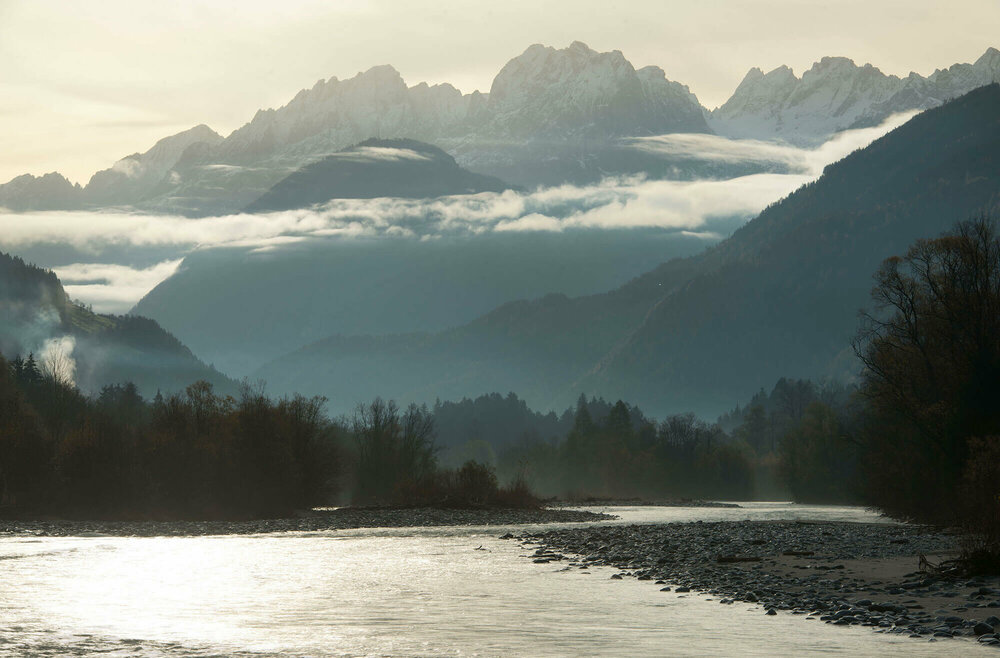 Die Isel macht ein paar Kurven. Im Hintergrund sieht man Berge, die von Nebelschwaden umhüllt sind.