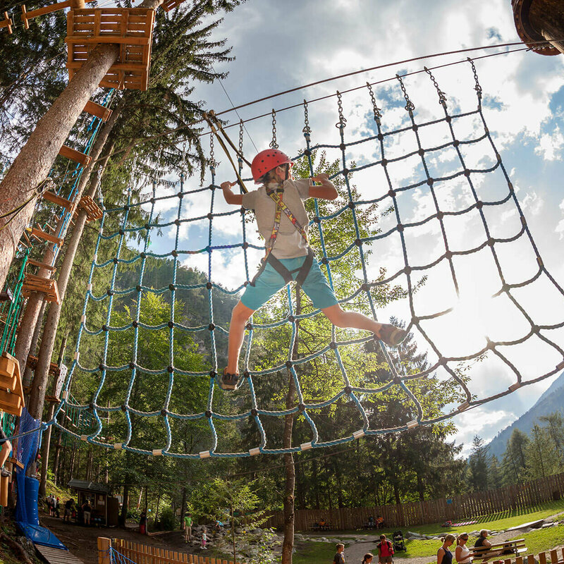 Kinder an einer Kletterstation im Hochseilgarten der Galitzenklamm bei sonnigem Wetter. Im Hintergrund spielen mehrere Personen am Wasserspielplatz. 