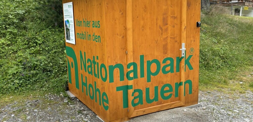 Eine hölzerne Hütte mit grüner Aufschrift "Nationalpark Hohe Tauern", in der sich der E-Scooter befindet.
