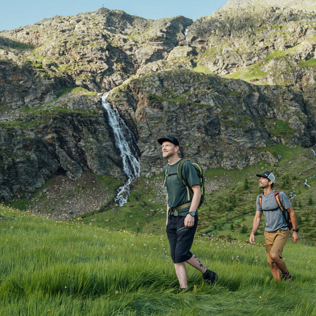 Zwei Männer wandern in Osttirols Bergen hinauf zum Geigensee durch hohes Bergwiesengras. Im Hintergrund ist ein kleiner Wasserfall zu sehen, der talwärts rinnt.