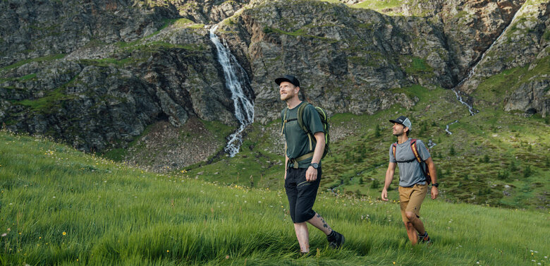 Zwei Männer wandern in Osttirols Bergen hinauf zum Geigensee durch hohes Bergwiesengras. Im Hintergrund ist ein kleiner Wasserfall zu sehen, der talwärts rinnt.