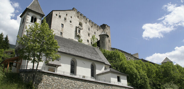 Die Burg Heinfels thront im Sommerlicht über der Landschaft, am Hang darunter liegt malerisch die kleine Kapelle.