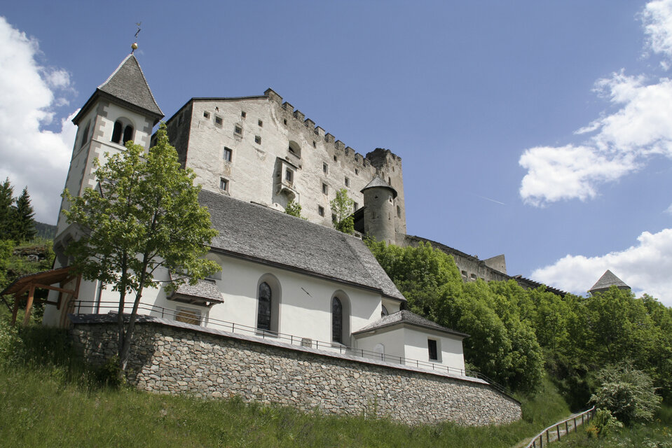 Die Burg Heinfels thront im Sommerlicht über der Landschaft, am Hang darunter liegt malerisch die kleine Kapelle.