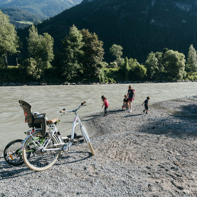 Eine Familie spielt am Ufer der Isel, im Vordergrund stehen zwei Räder eines in weiß und ein Kinderrad in giftgrün