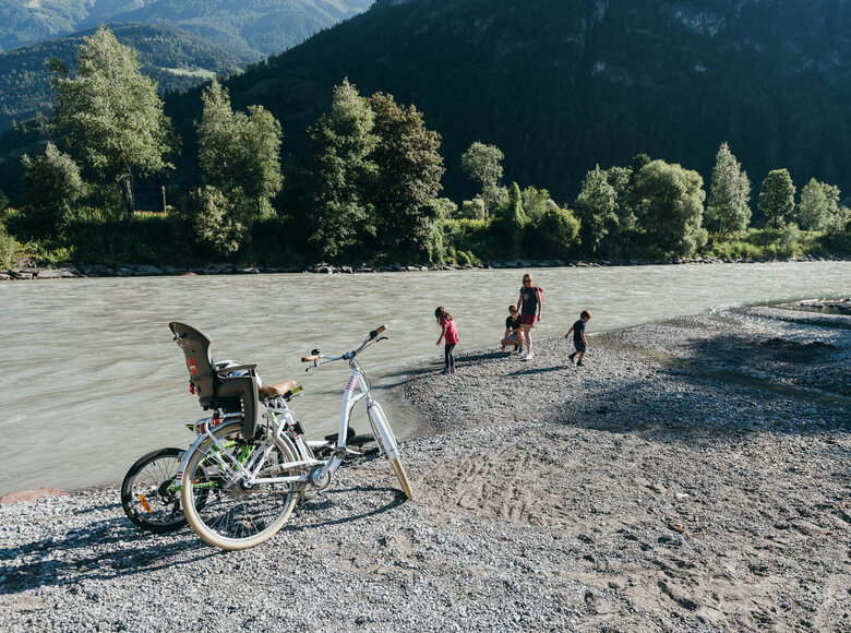 Eine Familie spielt am Ufer der Isel, im Vordergrund stehen zwei Räder eines in weiß und ein Kinderrad in giftgrün