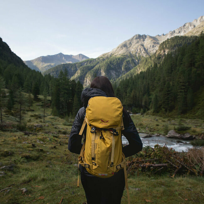 Wandern in Osttirol Wanderin mit gelbem Rucksack auf einer Lichtung in morgendlicher Stimmung kurz vor einem kleinen Gebirgsbach. Im Hintergrund strahlt die Morgensonne schon einen Bergrücken an.