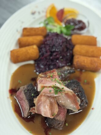 Das Bild zeigt einen Teller mit Hirschrücken, der ansprechend angerichtet serviert wird. Das Fleisch wird von Gemüse, einer Sauce und weiteren Beilagen begleitet.