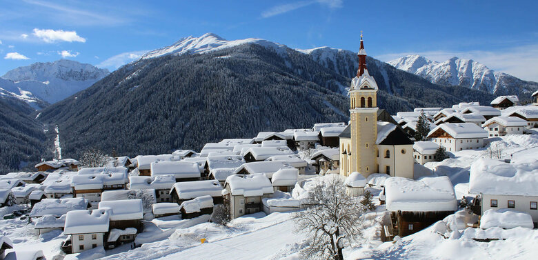  Viel Neuschnee liegt auf den Dächern des Dorfes mit der prägnanten Kirche im Vordergrund an einem fast wolkenlosen Wintertag. Im Hintergrund bewaldete und verschneite Bergrücken sowie tief verschneite Landschaft oberhalb der Baumgrenze mit zum Teil bizarr geformtem Bergkamm.
