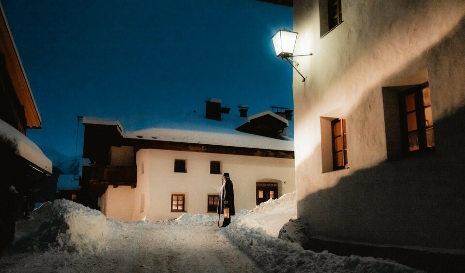 Der Nachtwächter ist mit einem Hut und einem Lodenmantel bekleidet und trägt einen Stock sowie eine Laterne mit sich durch die engen und verschneiten Gassen von Obertiliach.