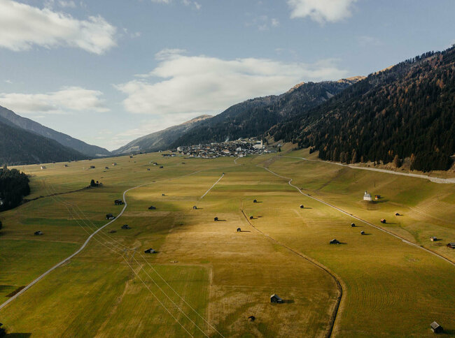 Herbstlich gebräunte Felder mit vielen kleinen Heuhütten und zwei am Feldrand stehenden Kapellen von der Luft aus betrachtet. Im Hintergrund ein kleines Bergdorf, das sich an den Bergfuß schmiegt und nochmals dahinter der V-formige Talabschluss und ein bewölkter Himmel.