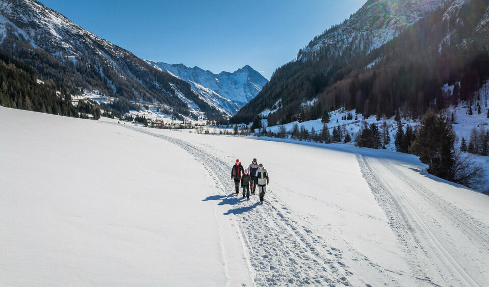 Eine Familie, bestehend aus zwei Elternteilen und zwei Kindern, wandert inmitten verschneiter Bergwelt. Die Sonne strahlt am wolkenlosen, blauen Himmel.