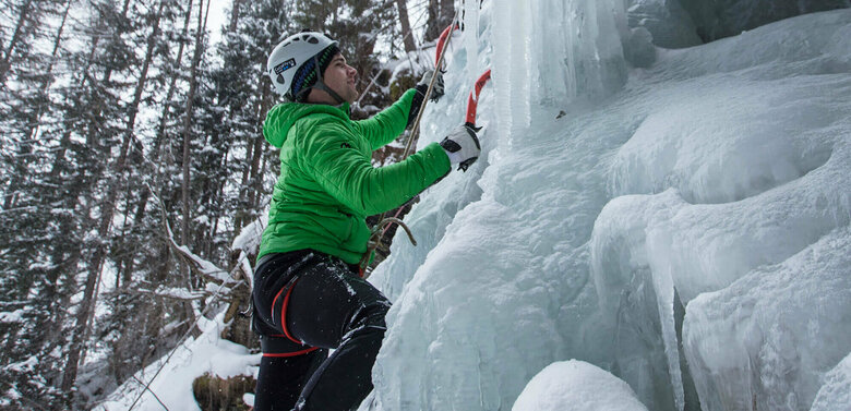 Eiskletterer in einer grünen Jacke, mit Pickel und Steigeisen auf einem zugefrorenen Wasserfall